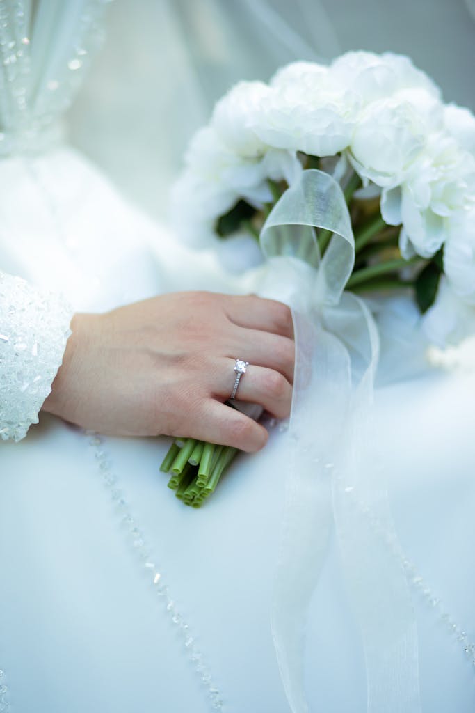 A bride's hand holding a white floral bouquet, showcasing an elegant wedding ring.